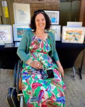 Rachel sitting in her wheelchair in a flowery dress smiling with her art work in the background