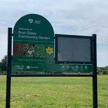 A green information sign for the organisation's community garden, featuring flower photographs and the logos of Wigan Council, Visit Wigan, and Greenheart, standing in an open grassy area under a cloudy sky.