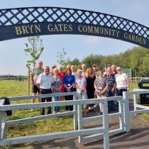 A group of around twenty adults smiling for a photo beneath the ornate arched entrance gate to the organisation's community garden on a sunny day, with an open green space and play equipment visible in the background.