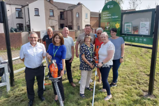 A group of around nine adults smiling together outdoors at the organisation's community garden, holding gardening tools including a hedge trimmer and strimmer, with the garden's welcome sign visible behind them.