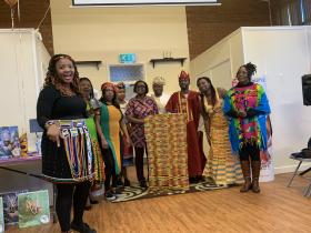 A group of people dressed in vibrant traditional African clothing and accessories performing or presenting at an indoor community event, with colourful kente cloth, beaded jewellery, and ceremonial robes on display. A "Islands of the Bahamas" promotional banner is visible in the background.