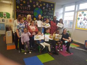 A group of children and two adults smiling at the camera inside Golborne Library, with several children proudly holding up their completed artwork or worksheets. A colourful space-themed display on the wall behind them reads "Visit Golborne Library - it's out of this world."