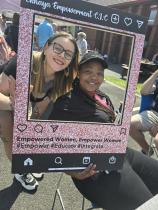 Two women smile together at an outdoor community event, posing behind a large decorative photo frame styled to look like an Instagram post, branded with the organisation's name and the tagline "Empowered women, empower women."