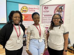 Three women wearing the organisation's lanyards smile together at an indoor event, standing in front of two promotional pull-up banners for the organisation and a partner organisation, Happy Smiles Training.