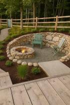 A circular stone-walled fire pit area with teal wooden chairs and a lit fire, surrounded by landscaped planting beds, a wooden decking area, and a rustic fence with trees in the background.
