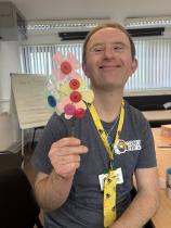 A portrait of a young male member of the organisation sits in a classroom and smiles while holding a handmade bunch of flowers made out of fabric. He wears a t-shirt with the logo of the organisation and a yellow lanyard around his neck with bees printed on it. 