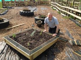 A smiling man kneels beside a newly built wooden raised garden bed with young plants in it, surrounded by reclaimed tyres used as planters and a woodchip surface on the farm.