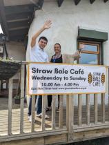 Two people wave cheerfully from the decking of the organisation's Straw Bale Café, standing behind a banner displaying the café's opening hours - Wednesday to Sunday, 10am to 4pm.