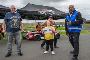Girl smiling with her hands in the air standing in front of a racing car with staff standing close smiling and cheering