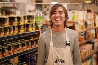Shopkeeper outside smiling in front of a sign reading Village Greens Coop